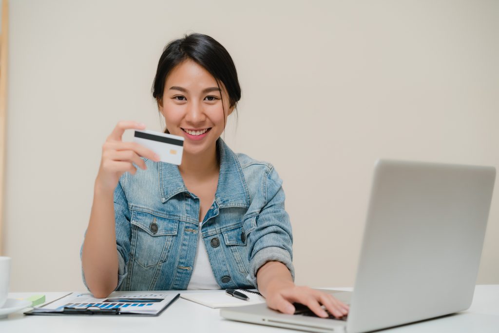 Beautiful smart business Asian woman using computer or laptop buying online shopping by credit card while wear smart casual sitting on desk in living room at home. Lifestyle woman at home concept.